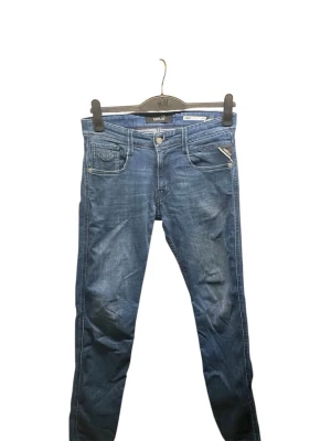 Blå slim fit jeans från Replay - Snygga blå jeans från Replay i modellen Anbass, slim fit. Klassisk femficksdesign med dragkedja och knapp. Jeansen har en cool tvätt och smal passform som sitter tajt längs benen. Perfekta för en trendig och avslappnad stil.