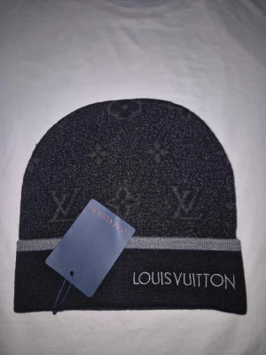 Svart mössa från Louis Vuitton - Snygg svart mössa från Louis Vuitton med diskret monogrammönster och grå kant. Märkets logga är broderad framtill på uppviket. Perfekt accessoar för dig som vill ha en exklusiv och stilren look.