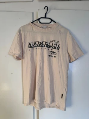 Beige Napapijri t-shirt med tryck - Snygg beige t-shirt från Napapijri med svart tryck framtill där det står 'Napapijri Geographic Outdoor Explorers'. Klassisk passform och rund hals. Perfekt till jeans eller shorts för en avslappnad look.
