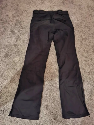 Svarta täckbyxor från Dope - Svarta täckbyxor från Didriksons med raka ben, breda bälteshällor och dubbla knappar i midjan. Byxorna är tillverkade i slitstarkt och vattenavvisande material, perfekta för vintersport och utomhusaktiviteter. Dragkedjor vid benslut för enkel påtagning över skor.