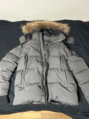 Grå dunjacka med pälskantad huva - Säljer en grå dunjacka från Canada Goose med avtagbar huva och pälskant. Jackan har ribbade muddar vid ärmsluten, två sidofickor och en patch med logga på ärmen. Perfekt för kalla vinterdagar tack vare tjockt dunfoder och vindtätt material.