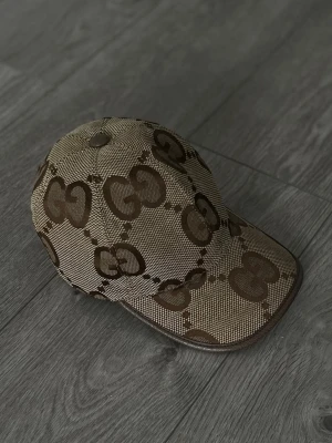 Beige och brun keps från Gucci - Säljer en beige och brun keps från Gucci med det klassiska GG-mönstret över hela kepsen. Kepsen har böjd skärm och är tillverkad i ett slitstarkt tyg med bruna detaljer längs kanterna. Perfekt för dig som vill ha en stilren och lyxig accessoar.