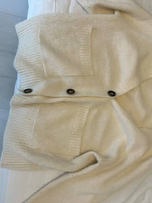 Beige kofta med stora knappar - Mysig beige kofta med stora mörka knappar framtill och två rejäla fickor. Koftan har ribbade muddar och är gjord i ett mjukt, stickat material som känns skönt mot huden. Perfekt att slänga på sig när det är lite kyligt ute.