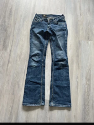 Blå bootcut jeans från Lee - Säljer ett par klassiska blå bootcut jeans från Lee med snygga slitningar och fem fickor. Jeansen har låg midja, dragkedja och knapp framtill samt Lee-lapp bak i midjan. Jeansen har en liten slitning längst ner på ena benet men annars väldigt bra skick💗längden är 33 och midjan 28😊