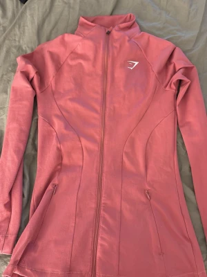 Rosa träningsjacka från Gymshark - Snygg rosa träningsjacka från Gymshark med hel dragkedja framtill och hög krage. Figurnära passform och långa ärmar. Tillverkad i ett stretchigt funktionsmaterial som passar perfekt till gymmet eller löprundan.