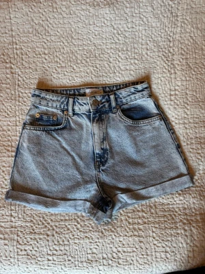 Högmidjade jeansshorts från NA-KD - Säljer ett par ljusblå högmidjade jeansshorts från NA-KD. Klassisk femficksmodell med uppvikta benslut och knappgylf. Perfekta till sommaren och lätta att matcha med olika stilar.