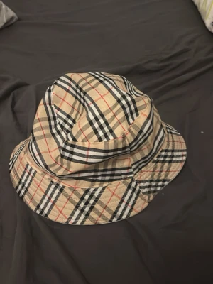 Beige bucket hat med rutigt mönster - Snygg bucket hat i beige med klassiskt rutigt mönster i svart, vitt och rött. Hatten har bred brätte och är tillverkad i ett mjukt tygmaterial. Perfekt accessoar för att lyfta din outfit.