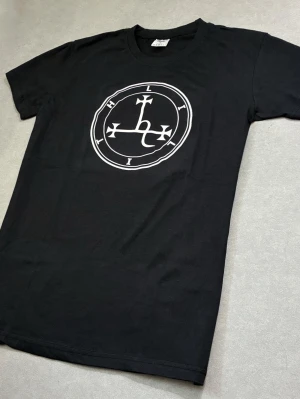 Lilith  t shirt och patch  - Snygg svart t-shirt med ett stort vitt tryck på bröstet i form av en cirkel med bokstäver och symboler. Klassisk rund hals och korta ärmar. Tillverkad i mjuk bomull som känns skön mot huden. Perfekt för dig som gillar grafiska prints.