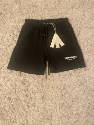 Svarta shorts Essentials Fear of God - Svarta shorts från Essentials Fear of God med vit logga på vänster ben. De har elastisk midja med snörning och är tillverkade i mjukt material, perfekta för chill dagar. Klassisk och enkel design med raka ben.