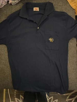 Marinblå pikétröja från Calvin Klein - Snygg marinblå pikétröja från Calvin Klein med ribbad struktur och klassisk krage. Tröjan har korta ärmar, knappslå framtill och en bröstficka med gul CK-logga broderad. Perfekt för en stilren och avslappnad look.