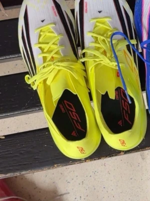 Adidas F50 gula fotbollsskor gräs - Snygga Adidas F50 fotbollsskor i neon-gult med svarta och röda detaljer. Skorna har en smal och aerodynamisk form, snörning framtill och ovandel i syntetmaterial. Perfekta för dig som vill sticka ut på planen och få bra grepp och känsla med bollen. Helt oanvänd!