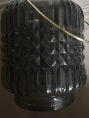 Mörkgrå glaslykta med handtag - Säljer en mörkgrå glaslykta med dekorativt mönster och guldfärgat metallhandtag. Perfekt för värmeljus eller små blockljus. Glaset har en räfflad och diamantmönstrad yta som ger ett snyggt sken när ljuset är tänt.