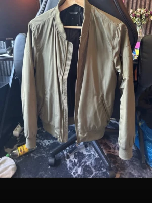 Olivgrön bomberjacka - Snygg olivgrön bomberjacka från The Black Label Collection i storlek S. Jackan har ribbade muddar vid ärmslut, krage och nederkant samt två sidofickor med dragkedja. Fodret är svart och quiltat för extra komfort.