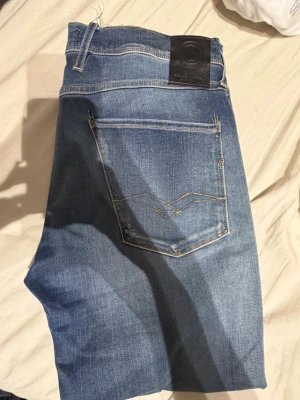 Replay anbass  - Säljer ett par ljusblå jeans med klassisk femficksdesign och en tydlig slitning vid knät samt råa, avklippta benslut. Jeansen har kontrastsömmar och en bakficka med dekorativ söm. Perfekta för en avslappnad och trendig look.