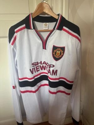 Manchester United retro långärmad tröja - Retro långärmad Manchester United-tröja i vitt med svarta och röda detaljer. Broderad klubbmärke på bröstet och SHARP VIEWCAM-tryck framtill. Klassisk krage med svart och röd kant. 