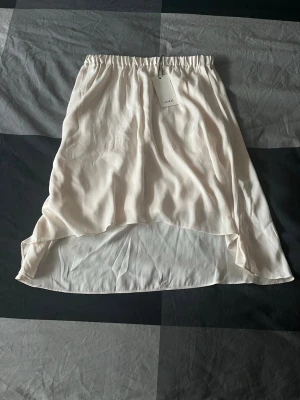 Ljusbeige omlottkjol i satinlook - Supersnygg ljusbeige kjol med omlottkänsla och elastisk midja. Kjolen har en flowy och lätt form, är längre bak och kortare fram vilket ger en cool asymmetrisk look. Materialet har en satinliknande finish som känns lyxig och trendig.