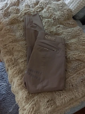 Beige ridbyxor med detaljer - Snygga beige ridbyxor med silikongrepp på insidan av benen och dekorativa nitar vid fickan. Byxorna har dragkedjeficka bak och smal passform. Perfekta för dig som vill ha både stil och funktion i stallet. Märke crw från hööks