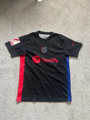 FC Barcelona svart matchtröja - Säljer en svart FC Barcelona matchtröja med röda och blå detaljer på sidorna. Tröjan har Spotify-logga på bröstet, klubbmärke och tryck med Lamine Yamal och nummer 19 på ryggen. Materialet är lätt och andas, perfekt för fotbollsträning.