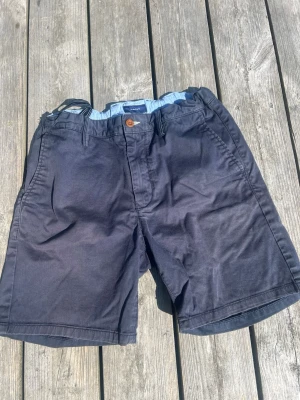 Mörkblå bomullsshorts från Gant, stl 158/164, 13-14y - Snygga mörkblå shorts från Gant i mjuk bomull. Klassisk design med knappstängning, bälteshällor och sidofickor. Perfekta för varma dagar och enkla att matcha med olika stilar.