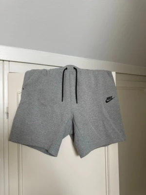 Gråa shorts från Nike med snörning - Gråa mjuka shorts från Nike med svart snörning i midjan och logga på vänster ben. Shortsen har fickor på sidorna och en bakficka med dragkedja. Perfekta för sommaren och avslappnade dagar.