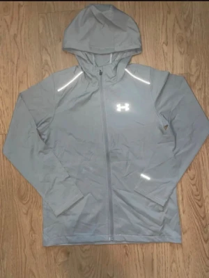 Ljusgrå hoodie från Under Armour - Snygg ljusgrå hoodie med dragkedja från Under Armour. Har reflekterande detaljer på axlar och ärm, samt en klassisk huva. Perfekt för träning eller chill. Tillverkad i ett lätt och funktionellt material.