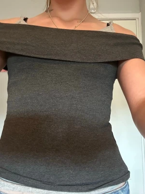 Mörkgrå offshoulder topp - Snygg mörkgrå offshoulder topp med bred krage som vilar över axlarna. Toppen är tight och har en enkel, stilren design utan mönster eller detaljer. Perfekt att matcha med jeans eller kjol för en trendig look.