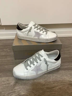Golden Goose skor - Snygga vita sneakers med grå mockastjärna på sidan, grå skosnören och svart detalj på hälen. Skorna har en klassisk låg profil och sula med mönster. Perfekta för en trendig och avslappnad stil.