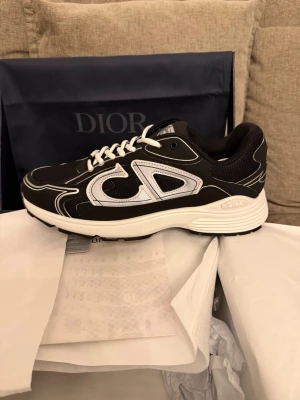 Svarta  sneakers med vit sula - Snygga svarta sneakers  med vit och svart sula samt vita detaljer och snörning. Skon har en stor vit logga på sidan och är tillverkad i en mix av textil och läder. Perfekt för dig som vill ha en exklusiv och trendig look.