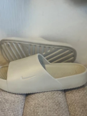 Beige tofflor från Nike - Säljer ett par stilrena beige tofflor från Nike med enkel design och diskret logga på sidan. Tillverkade i ett mjukt och flexibelt material med räfflad innersula för extra komfort. Perfekta för att glida runt hemma eller på stranden.