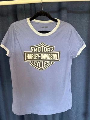 Ljusblå Harley-Davidson t-shirt - Snygg ljusblå t-shirt från Harley-Davidson med klassisk logga framtill. T-shirten har vita kontrastdetaljer vid halsringning och ärmslut. Perfekt för dig som vill ha en cool och avslappnad stil. Sitter mer som en Medium! 