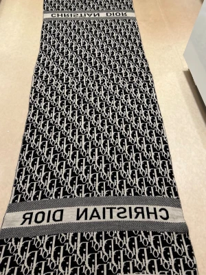 Svartvit halsduk från Christian Dior - Snygg svartvit halsduk från Christian Dior med ikoniskt logomönster och breda ränder med märkets namn. Halsduken är lång och rektangulär, perfekt att svepa flera varv. Materialet ser ut att vara mjukt och stickat, vilket ger en lyxig känsla.