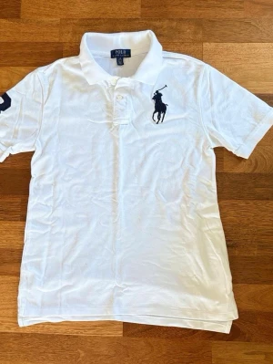 Vit pikétopp från Polo Ralph Lauren - Vit pikétopp med klassisk krage och korta ärmar från Polo Ralph Lauren. Toppen har broderad mörkblå logga på bröstet och siffrad detalj på ärmen. Tillverkad i mjuk bomull för en bekväm och stilren look.