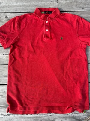 Morris Piké Röd - Säljer en klassisk röd pikétröja med krage och två knappar framtill. Tröjan har korta ärmar och en liten broderad logga på bröstet. Perfekt för en avslappnad men ändå stilren look.
