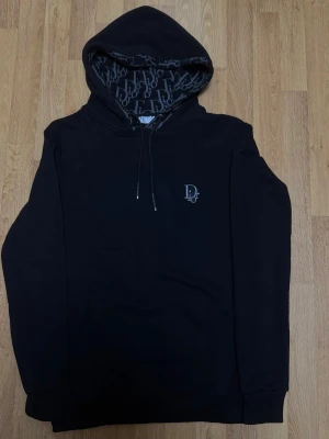 CD hoodie - Size M  - Stilren och bekväm hoodie i svart med broderad logga på bröstet och monogrammönster på insidan av huvan. Hoodien är i mycket bra skick och har inga fel. Storlek M (liten i storlek)  Rabatt vid köp av flera plagg!