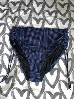 Bikiniunderdel - Snygg bikiniunderdel i mörkblått med rutigt mönster och justerbar snörning i sidorna. Hög midja och bekväm passform, perfekt för strandhäng eller poolen. Materialet känns mjukt och följsamt mot huden. Helt ny, men tyvärr har hygienskyddet i skrevet lossnat då denna legat i förrådet.