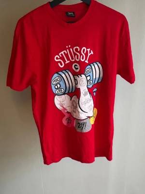 Röd Stüssy t-shirt med tryck - Röd t-shirt från Stüssy med färgglatt tryck framtill som föreställer en arm som lyfter hantlar. T-shirten har rund halsringning och korta ärmar. Perfekt för dig som gillar streetwear och vill sticka ut med en unik design.