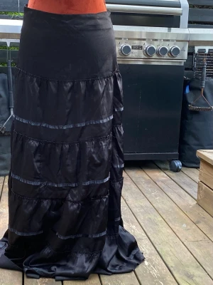 Svart långkjol i satin med volanger 36 - Lång svart kjol med volanger och banddetaljer. Kjolen har i sidan för stängning. Längd: 96 cm Midja: 76 cm Uppskattad storlek S  Inga bilder när den sitter på mig! Katter finns i hemmet!