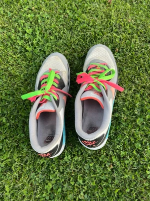 Nike sneakers med färgglada snören - Säljer ett par gråa Nike sneakers med mesh- och syntetmaterial. Skorna har coola kontrasterande skosnören i neonrosa och neongrönt samt detaljer i svart och vitt. Låg modell med klassisk Nike-logga på plösen och hälen. Perfekta för dig som vill sticka ut!