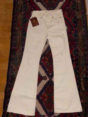 Vita utsvängda jeans med tagg - Snygga vita jeans med utsvängda ben och klassisk femficksmodell. Jeansen har låg midja och lätt slitna detaljer framtill. Perfekta för dig som gillar retrostil och vill sticka ut med en cool siluett.