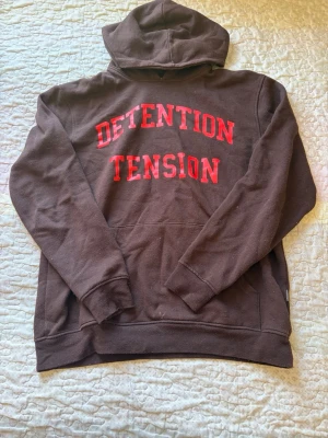 Brun hoodie med röd text DETENTION - Säljer en brun hoodie med huva och stor ficka framtill. På bröstet står det 'DETENTION TENSION' i stora röda bokstäver. Hoodien har en klassisk passform och är gjord i mjukt sweatshirtmaterial, perfekt för en avslappnad stil.