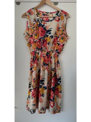 Blommig ärmlös sommarklänning, storlek M - Härlig blommig klänning i storlek M med elastisk midja och ärmlös design. Klänningen har en ljus cremefärgad bas med färgglatt mönster med stora blommor i rosa, gult och blått. Perfekt för varma dagar och har en avslappnad passform som ger en luftig känsla. Mycket gott skick, som ny! 