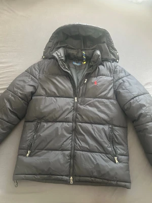 Svart dunjacka från Polo Ralph Lauren - Snygg och stilren dunjacka från Polo Ralph Lauren i svart färg med huva och två dragkedjeförsedda fickor. Jackan har det klassiska röda Polo-logotypen broderad på bröstet och quiltad design. Perfekt för kalla vinterdagar och riktigt skön att bära.