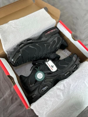 Svarta Nike TN  Air Max Plus sneakers - Säljer ett par svarta Nike Air Max Plus sneakers med klassisk TN-design. Skorna har snörning, mesh- och syntetmaterial samt svarta detaljer på sulan. Perfekta för dig som gillar streetwear och vill ha en clean look.