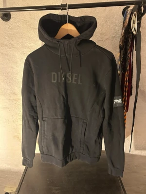 Svart hoodie från Diesel med logga - Snygg svart hoodie från Diesel med stor logga på bröstet och en patch med Diesel-märke på ärmen. Klassisk känguruficka framtill och justerbar huva med dragsko. Perfekt för en avslappnad och trendig look.