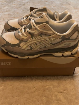 Asics vita och gråa sneakers - Snygga sneakers från Asics i vitt, beige och ljusgrått med mesh- och mockadetaljer. Skorna har klassisk låg profil, tydliga Asics-loggor på sidan och en chunky sula för extra komfort. Perfekta för dig som gillar stilrena och sportiga vibes. 