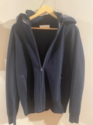 | Sayless | Mörkblå stickad cardigan  - Aldrig använd! Mörkblå stickad cardigan från Sayless med dragkedja framtill och två fickor. Huvan har ett quiltat, glansigt tyg i grått som ger en schysst kontrast. Perfekt för dig som gillar en avslappnad men ändå stilren look.