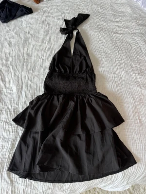 Svart halterneck volangklänning - Säljer en svart kort klänning med halterneck och knyt i nacken. Klänningen har smock i midjan och dubbla volanger i kjolen för en extra flowy look. Perfekt för dig som gillar en feminin och trendig stil.