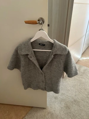 Grå kortärmad stickad kofta - Kortärmad grå stickad kofta med krage och knappar framtill. Koftan har en avslappnad passform och ribbade kanter vid ärmslut och nederkant. Perfekt att slänga över en topp för en chill look!! 👀💞(Andvänt 1 gång)