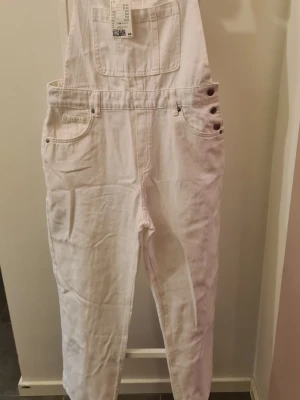 Vit jeansbyxdress med ficko prislapp kvar. - Säljer en vit jeansbyxdress med klassisk hängselmodell. Byxdressen har flera fickor fram och bak samt knappdetaljer på sidan. Tillverkad i denim med raka ben och justerbara axelband. Perfekt för en avslappnad och trendig look.