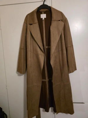 Ljusbrun trenchcoat med bälte - Snygg ljusbrun trenchcoat i mockaimitation med långa ärmar och bred krage. Kappan har ett matchande bälte i midjan och djupa fickor på sidorna. Perfekt för dig som vill ha en stilren och trendig look till hösten.